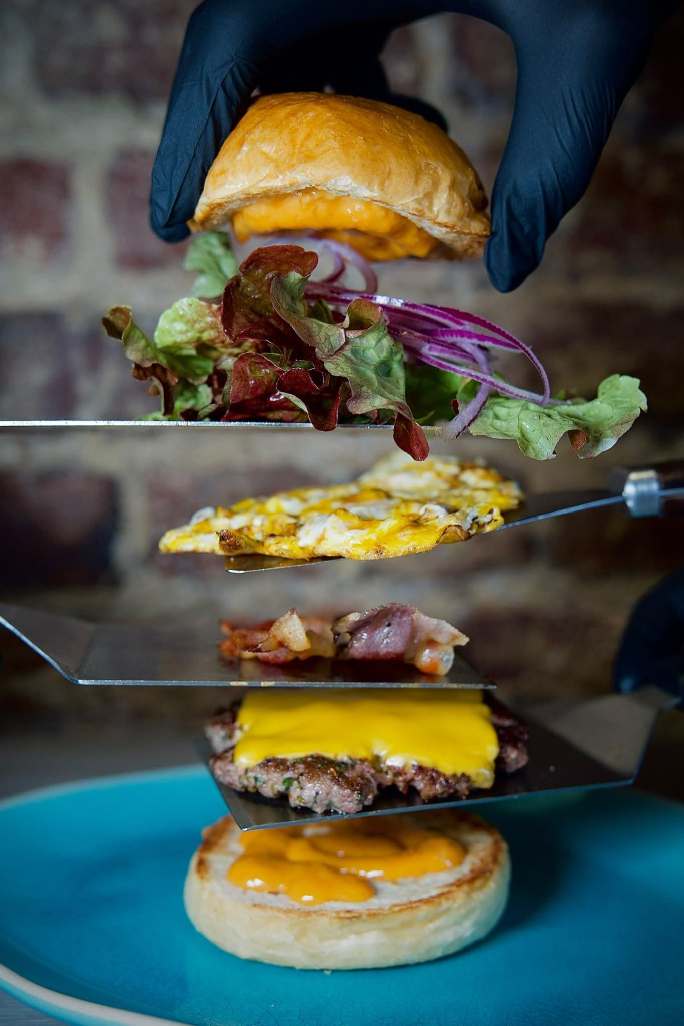 Un burger tenu ouvert sur des spatules devant le mur de briques de la salle : le pain du dessus, la salade et l’oignon rouge, l'œuf, le lard, le steak au cheddar, et le pain du dessous nappé de sauce.