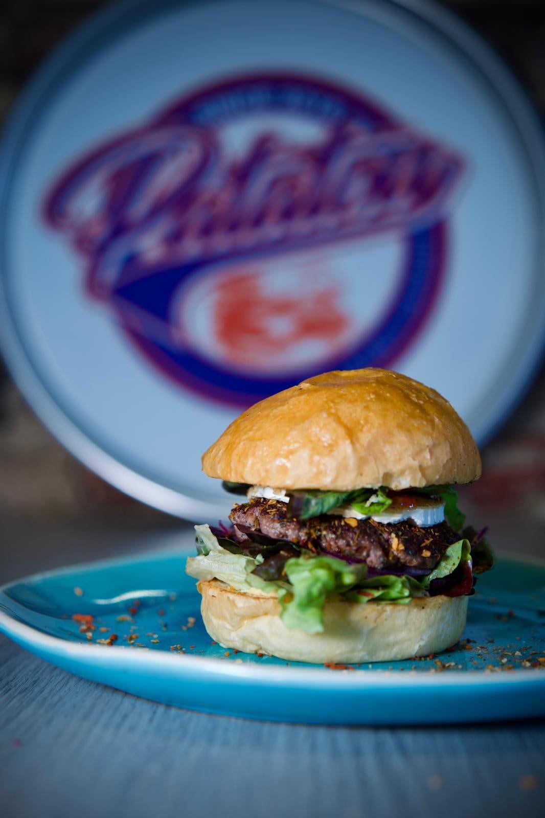 Un burger servi sur une assiette devant le logo de la friterie peint au mur.