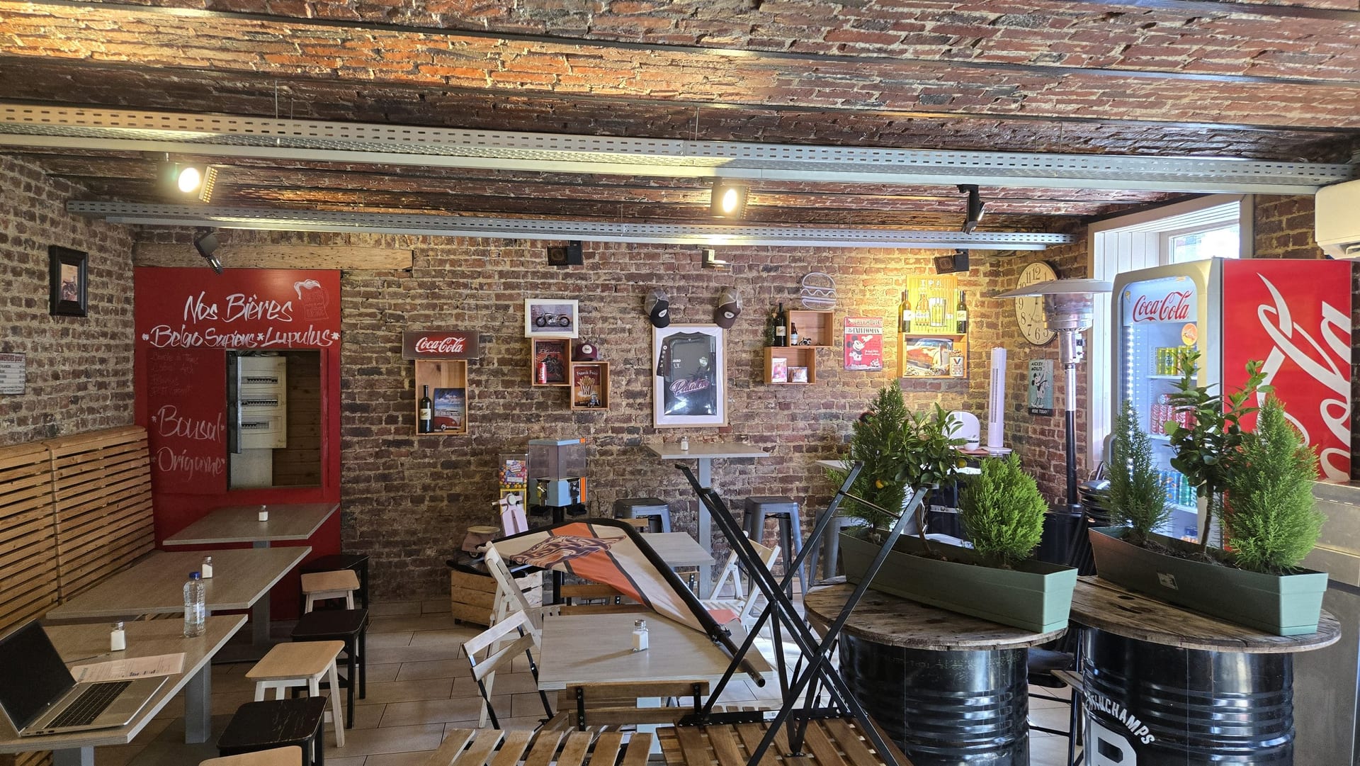 La salle de la friterie : murs de briques, poutres apparentes, tables et tabourets en métal.